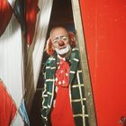 Ein Clown mit roter Nase und orangenen Haaren schaut durch einen Vorhang. Er trägt einen karierten Mantel und eine gepunktete Fliege.