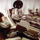 Menschen in futuristischen Sesseln tragen identische Raumanzüge in einem Raumschiff. Links unten DEFA-Logo.