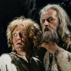 Zwei Männer mit besorgten Blicken. Der eine hat blondes, lockiges Haar, der andere graues Haar und trägt Ketten.