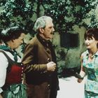 Vor einem Altbau stehen drei Personen: Eine Frau in Tracht, ein älterer Mann in Jacke, eine Frau in blauem Sommerkleid.