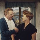 Ein Mann und eine Frau in einer rustikalen Stube stehen voreinander. Draußen sieht man Berge durch das Fenster.