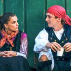 Ein Mann und eine Frau tragen traditionelle Kleidung und sitzen vor einer grünen Wand, in ein Gespräch vertieft.