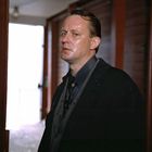 Es ist eine Person in einem dunklen Anzug in einem Holzgang, schaut ernst nach vorne.
