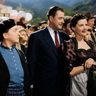 „Bau-Bel“ ist textlich sichtbar. Eine Gruppe von Menschen auf einem Fest, ein Mann in Anzug, daneben zwei Frauen in traditionellen Kleidern.