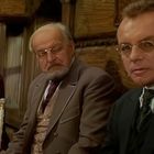 Drei ältere Männer in Anzügen sitzen zusammen, schauen ernst. Sie tragen Brillen, die Umgebung wirkt hölzern-gemütlich.