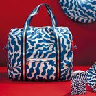 Kos Logo auf rotem Etikett. Große Tasche mit blau-weißem Muster, passende Notizbücher daneben. Hintergrund rot.