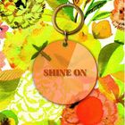 „SHINE ON“ steht auf einem runden Anhänger. Buntes, blumiges Hintergrundmuster mit leuchtenden Farben.