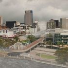 In der Mitte Gebäude mit Schildern und Logos; im Hintergrund Hochhäuser, ein Fluss im Vordergrund und Wolken darüber.