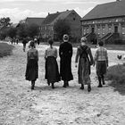 Mehrere Frauen gehen auf einem Dorfweg, umgeben von alten Backsteinhäusern; Hühner laufen frei herum.