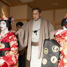 Zwei Personen in traditionellem japanischem Gewand; eine dritte steht zwischen ihnen. Alle tragen kunstvolle Kopfbedeckungen.