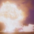 Es gibt keinen Text. Eine große Explosion erhellt den Nachthimmel, und Rauch steigt vom Boden auf.