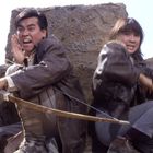 Es gibt keinen sichtbaren Text. Ein Mann und eine Frau in Lederjacken ducken sich hinter einem Felsen, der Mann hält einen Bogen.