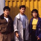 Drei Menschen vor gelber Wand. Eine Frau in blauer Jacke, ein Mann mit Waffe in grauem Mantel, ein Mann in brauner Jacke.