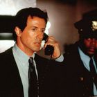 Ein Mann in Anzug spricht in ein schwarzes Telefon. Neben ihm steht ein Polizist in Uniform.