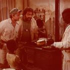 Vier Männer in einer rustikalen Küche, zwei in Safari-Kleidung, im Gespräch mit einem Koch an einem Herd.