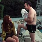 Ein Mann in Badehose plaudert neben einem Pool mit einer Frau, die auf einer Kante sitzt. Eine Rutsche ist im Hintergrund.