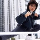 Ein Mensch in Kampfpose vor hohen, grauen Gebäuden. Er trägt eine dunkle Jacke, Jeans und weiße Turnschuhe.