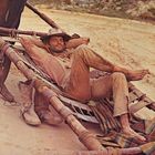 Ein Mann liegt entspannt in einem improvisierten Karren, der von einem Pferd gezogen wird, und trägt einen Hut.