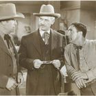 Drei Männer in Anzügen und Hüten in einem Raum; einer hält ein Dokument. Vintage-Western-Stil.