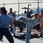 Ein Mann im grauen Trainingsanzug tritt mit einem hohen Kick gegen einen anderen. Andere Männer schauen zu. Sportgeräte im Hintergrund.