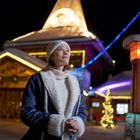 Eine Frau steht warm angezogen vor einem beleuchteten Holzgebäude mit Weihnachtsbaum und Lichterketten.