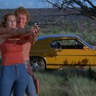 Eine Frau in einem roten Top hält mit Unterstützung eines Mannes eine Pistole. Im Hintergrund ein gelbes Auto in der Natur.