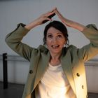 Eine Frau in einem grünen Blazer lächelt und formt ein Dreieck über ihrem Kopf mit ihren Händen.