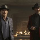 Zwei Männer mit Hüten, eleganten Anzügen, Feuerstelle im Hintergrund, einer hält ein Glas.