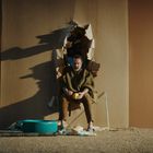 Ein Mann sitzt in einem Loch in der Wand, umgeben von Kartonresten. Vor ihm ein kleiner Pool und eine Krücke im Sand.