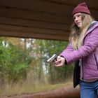 Eine Frau mit violetter Jacke und Mütze zielt mit einer Pistole in einem bewaldeten Gebiet unter einer Brücke.