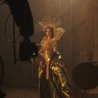 Kein sichtbarer Text. Eine Person in einem goldenen, eleganten Kostüm mit strahlender Kopfbedeckung in einer dramatischen Pose.