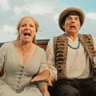 Ein Mann und eine Frau sitzen nebeneinander, beide mit schockierten Gesichtsausdrücken, blauem Himmel im Hintergrund.