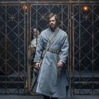 Ein Mann in grauem Gewand steht vor einem Metallgitter, ein weiterer sieht durch die Gitterstäbe. Die Umgebung wirkt düster.