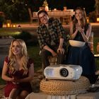 Eine Gruppe lachender Menschen sitzt draußen im Garten mit einem Projektor und Kerzenlichtern umgeben.