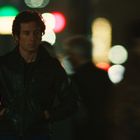 Ein Mann in dunkler Lederjacke steht nachdenklich in einer verschwommenen, belebten Straßenkulisse mit bunten Lichtern.