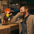 Ein Mann lächelt eine kleine animierte Figur an, die auf einem Spielzeugpferd sitzt. Werkstattumgebung.