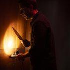 Ein Mann hält ein Messer und eine Taschenlampe vor einer Tür. Er wirkt angespannt und ist von warmem Licht beleuchtet.