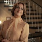 Eine lächelnde Frau in beigem Blazer mit Schal vor einer stilvollen Treppe im Hintergrund.