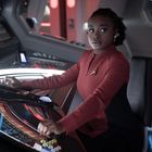 Eine Frau in roter Uniform sitzt in einer Hightech-Kontrollzentrale und bedient ein futuristisches Panel.