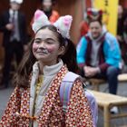 Ein lächelndes Kind in buntem Kostüm mit Hasenohren und Katzengesicht ist inmitten von bunt gekleideten Menschen.