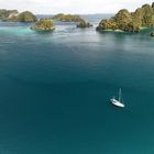 Kein Text enthalten. Ein weißes Segelboot auf türkisfarbenem Wasser, umgeben von üppigen, bewachsenen Inseln.