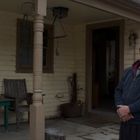 Eine verlassene Veranda mit Holzbank, Tisch, geschnitzter Ente und einem Mann in schwarzer Kapuzenjacke. Schneereste.