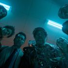 Sechs Personen schauen von oben herab, beleuchtet von blauem Licht. Eine Person trägt einen Anzug aus Drähten.