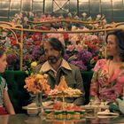 Drei Personen sitzen an einem Tisch inmitten eines blühenden, dekorativen Gartens voller bunter Blumen.