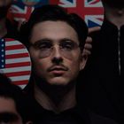 Eine Person mit Brille, umgeben von Schildern mit britischer und amerikanischer Flagge.