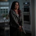 Eine Frau mit besorgtem Gesicht hält eine Pistole in einer Küche und trägt eine graue Jacke und ein rotes Kapuzenfutter.