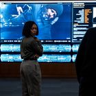 Daten auf Bildschirmen: "James Dormann", technische Details. Frau in grauer Kleidung, großer Monitor mit Video von schlafendem Mann.