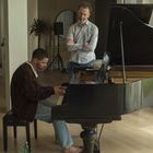 Ein Mann spielt Klavier, ein anderer beobachtet. Im Hintergrund stehen Personen in einem Raum mit Pflanzen und Fenstern.
