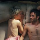 Keine Texte zu sehen. Zwei Menschen liegen nebeneinander im Bett, sie sehen sich an und wirken entspannt und vertraut.