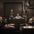 Eine Szene im Gerichtssaal mit einem Mann im Anzug am Rednerpult, umgeben von Soldaten. Zuschauer tragen Kopfhörer.